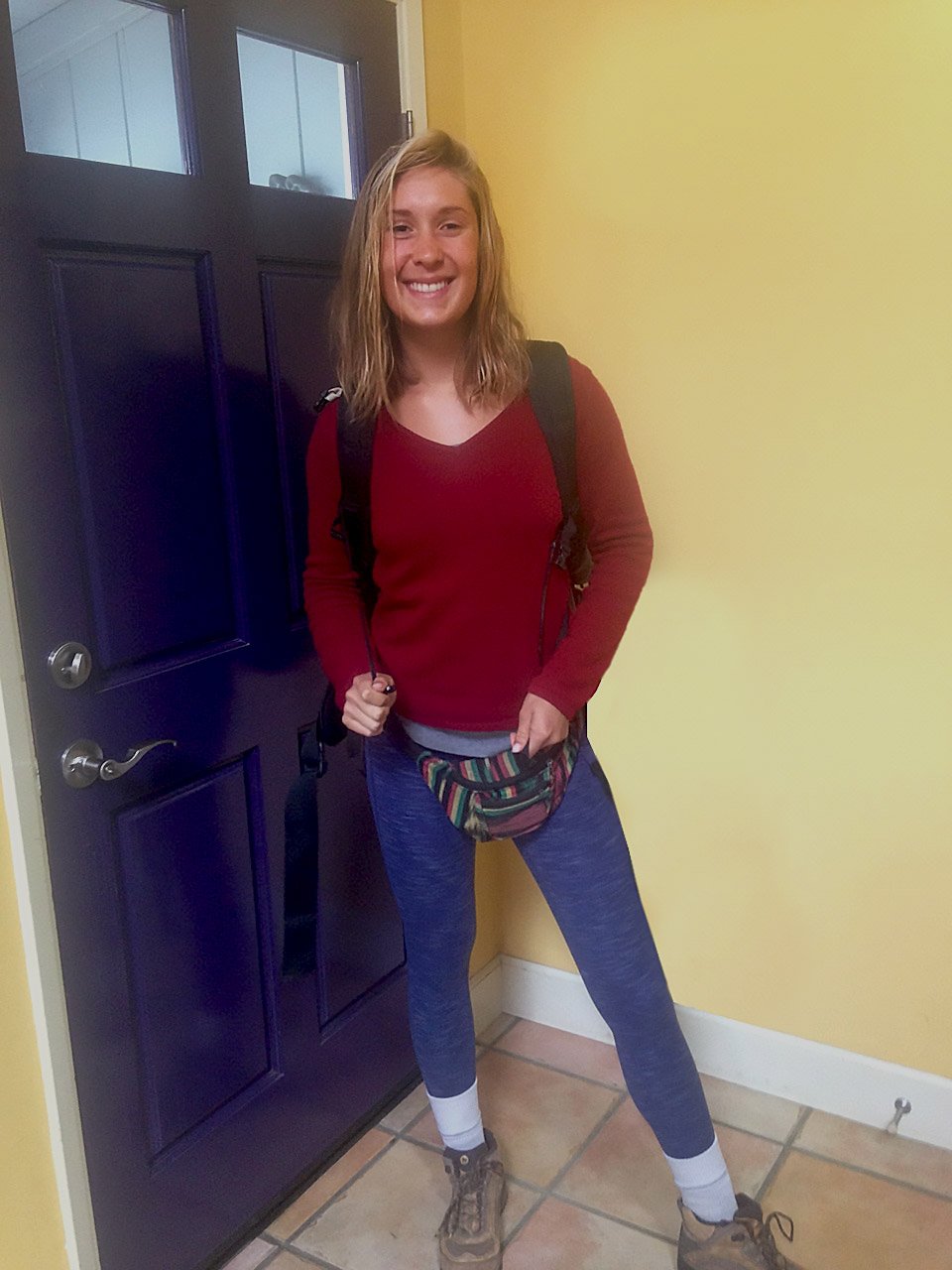 A photo of a young woman is standing indoors in front of a purple door, smiling at the camera. She is dressed in hiking attire, wearing a red long-sleeve shirt, blue leggings, hiking boots, and a colorful fanny pack. She also has a backpack on and appears ready for an outdoor adventure. The room has yellow walls and tile flooring, creating a bright, warm atmosphere.