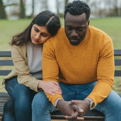 Photo of a couple is sitting on a bench outdoors, both looking down with a somber expression. The woman rests her head on the man's shoulder and has her hand gently on his arm, offering comfort. The man, wearing a mustard yellow sweater, appears deep in thought while holding his hands together. The woman, dressed in a beige cardigan and gray top, shows support as they sit quietly together in a peaceful park setting.