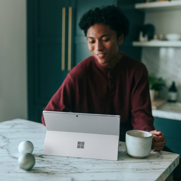 A photo of a person sitting at a table, using a tablet. They are holding a mug, likely enjoying a warm drink, while focused on the screen. The person is wearing a dark red sweater. The atmosphere appears relaxed and cozy, suggesting the person may be on a zoom therapy call.