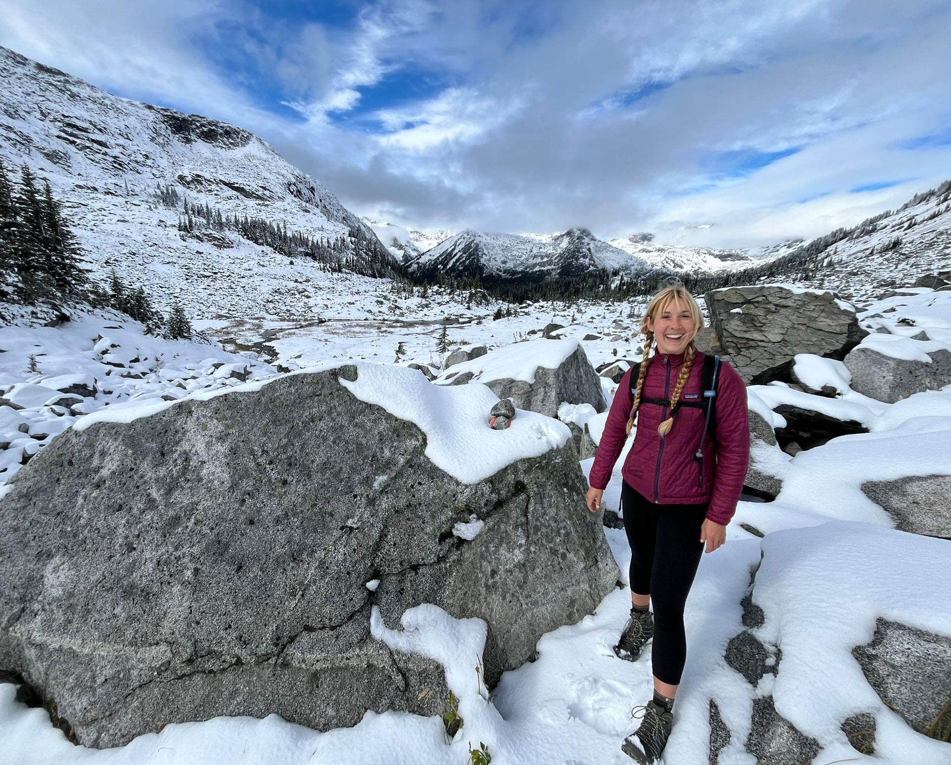 Eco-therapist-Sienna-Forest-standing-on-a-snowy-mountain-trail-surrounded-by-large-boulders-and-snow-covered-peaks Photo of Eco-therapist Sienna Forest standing on a snowy mountain trail, surrounded by large boulders and snow-covered peaks. She is wearing a maroon jacket, black leggings, and hiking boots under a bright, partly cloudy sky.