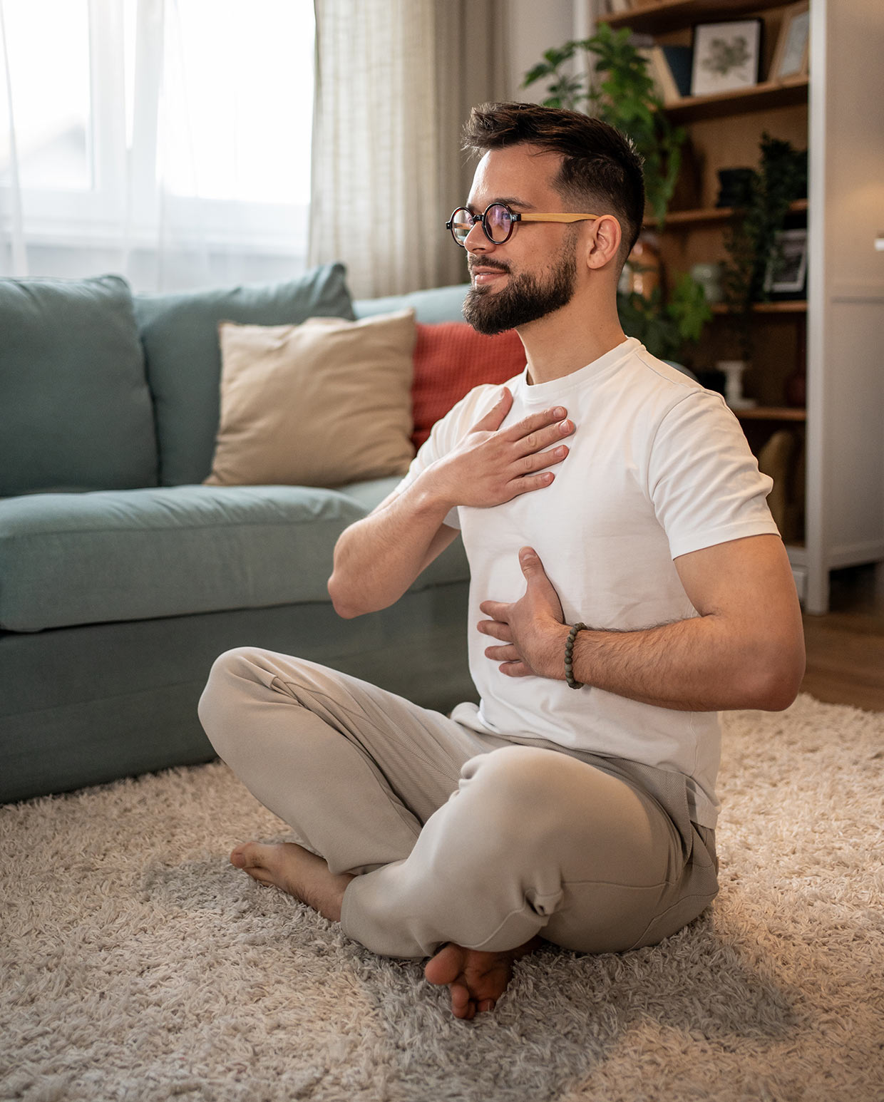 man-sitting-cross-legged-on-rug-in-cozy-room-practicing-mindful-breathing-with-one-hand-on-his-chest-and-the-other-on-his-stomach Photo of a man sitting cross-legged on a rug in a cozy room, practicing mindful breathing with one hand on his chest and the other on his stomach. He wears a calm expression on his face.