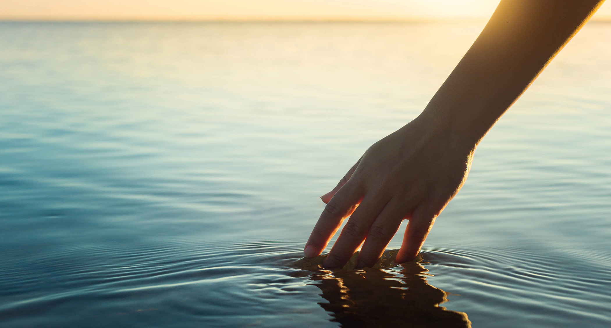 Photo of a hand gently touches the surface of calm water at sunset, creating soft ripples that reflect the golden light.