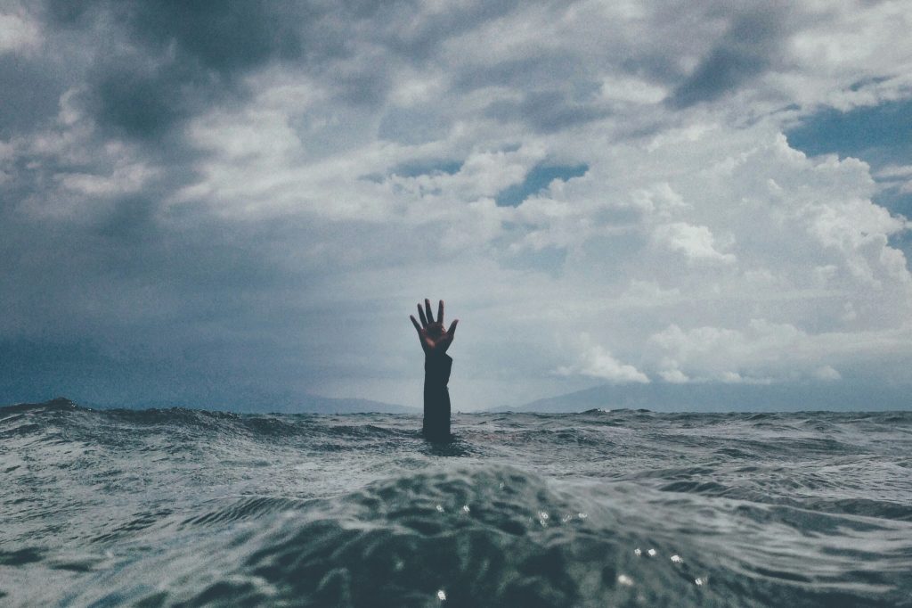 Photo of a single hand reaches out of choppy ocean waves beneath a stormy sky, symbolizing the experience of emotional overwhelm and dysregulation. The image evokes the feeling of being submerged in intensity—yet also the instinct to reach for air, balance, and self-support amid chaos.