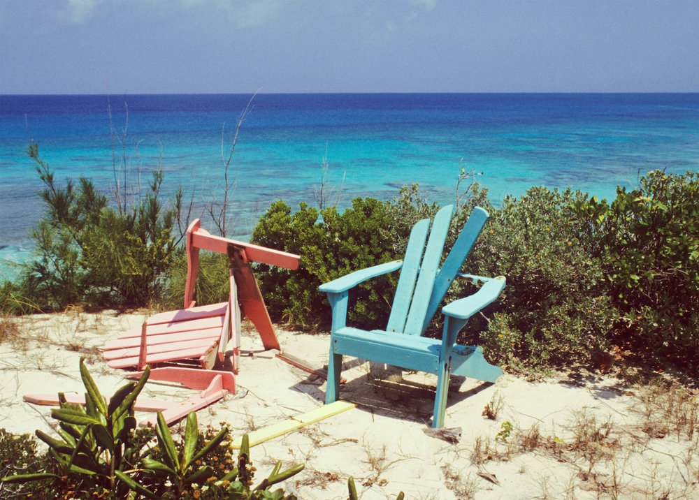 A photo of two colorful, broken Adirondack chairs sitting on the edge of a cliff in the sunshine.