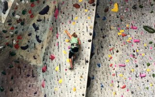 A photo of a person is climbing an indoor rock wall using colorful hand and foot holds. The wall features a variety of routes with different difficulty levels, indicated by the holds of various shapes and sizes. The climber, wearing a green tank top and black pants, is secured by a harness attached to a rope for safety. The wall has a steep incline, and the climber is midway up, reaching for the next hold.