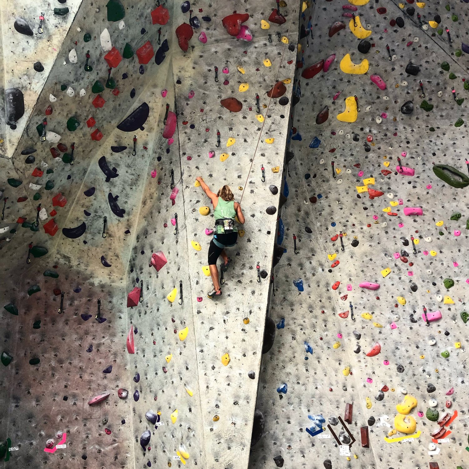 A photo of a person is climbing an indoor rock wall using colorful hand and foot holds. The wall features a variety of routes with different difficulty levels, indicated by the holds of various shapes and sizes. The climber, wearing a green tank top and black pants, is secured by a harness attached to a rope for safety. The wall has a steep incline, and the climber is midway up, reaching for the next hold.