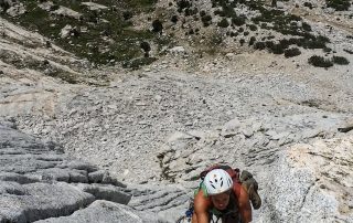 A photo of a climber is scaling a steep, rocky mountain face in an outdoor setting. The person, wearing a white helmet and a green tank top, uses both hands to grip the jagged rock as they ascend. The terrain below is a mix of rugged, rocky ground with sparse vegetation. The climber is secured by a rope and appears focused on the challenging ascent.