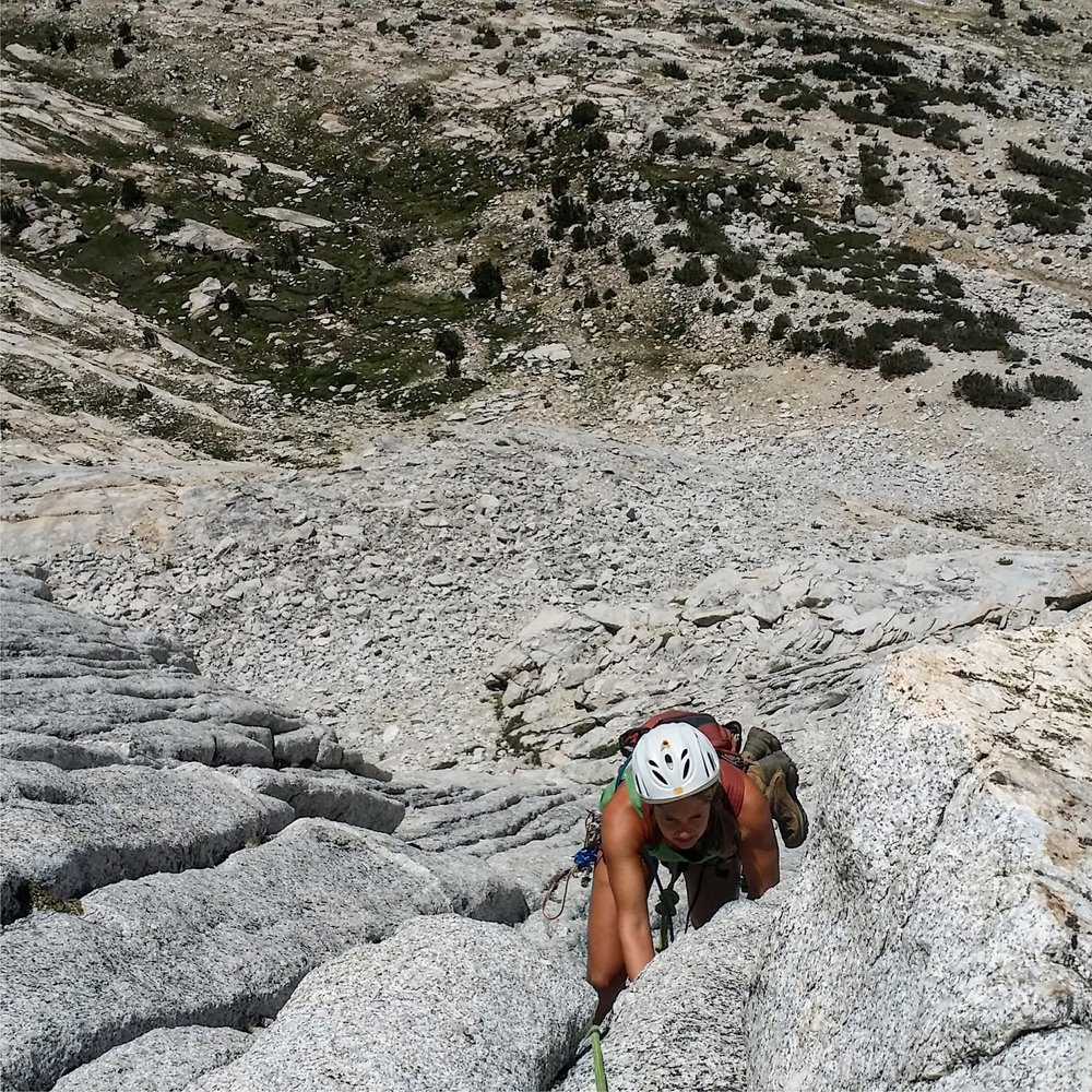 A photo of a climber is scaling a steep, rocky mountain face in an outdoor setting. The person, wearing a white helmet and a green tank top, uses both hands to grip the jagged rock as they ascend. The terrain below is a mix of rugged, rocky ground with sparse vegetation. The climber is secured by a rope and appears focused on the challenging ascent.