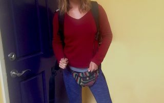 A photo of a young woman is standing indoors in front of a purple door, smiling at the camera. She is dressed in hiking attire, wearing a red long-sleeve shirt, blue leggings, hiking boots, and a colorful fanny pack. She also has a backpack on and appears ready for an outdoor adventure. The room has yellow walls and tile flooring, creating a bright, warm atmosphere.