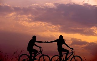 Two cyclists silhouetted at sunset, reaching out to hold hands as they ride side by side. The scene captures connection, togetherness, and balance, framed by a dramatic sky with soft, glowing clouds.