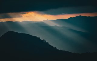 Photo of sunlight breaking through dark storm clouds over layered mountain ridges, symbolizing the movement between emotional intensity and calm. The image evokes the natural rhythm of regulation—how moments of darkness and light coexist, reminding us that dysregulation isn’t something to fix, but a wave to breathe through and find balance within.