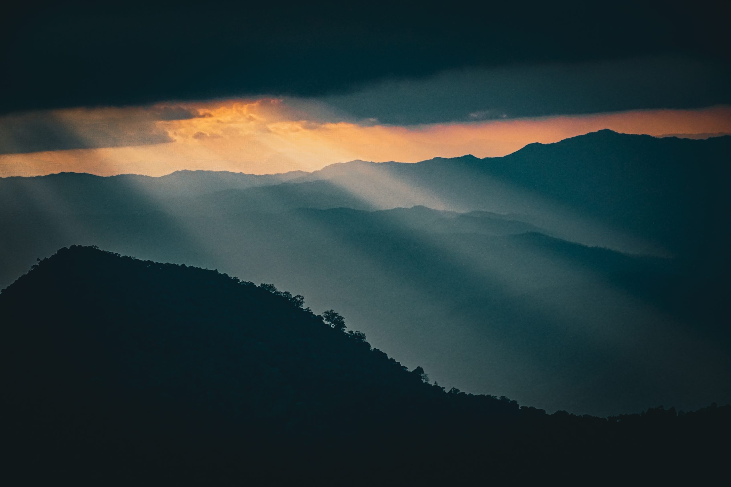Photo of sunlight breaking through dark storm clouds over layered mountain ridges, symbolizing the movement between emotional intensity and calm. The image evokes the natural rhythm of regulation—how moments of darkness and light coexist, reminding us that dysregulation isn’t something to fix, but a wave to breathe through and find balance within.